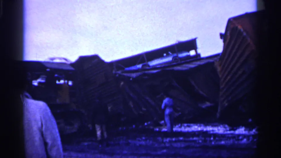 1961 Minnesota 8mm frame of overturned train cars in a snowy field, with adult bystanders partly visible in the foreground and near the wreckage.