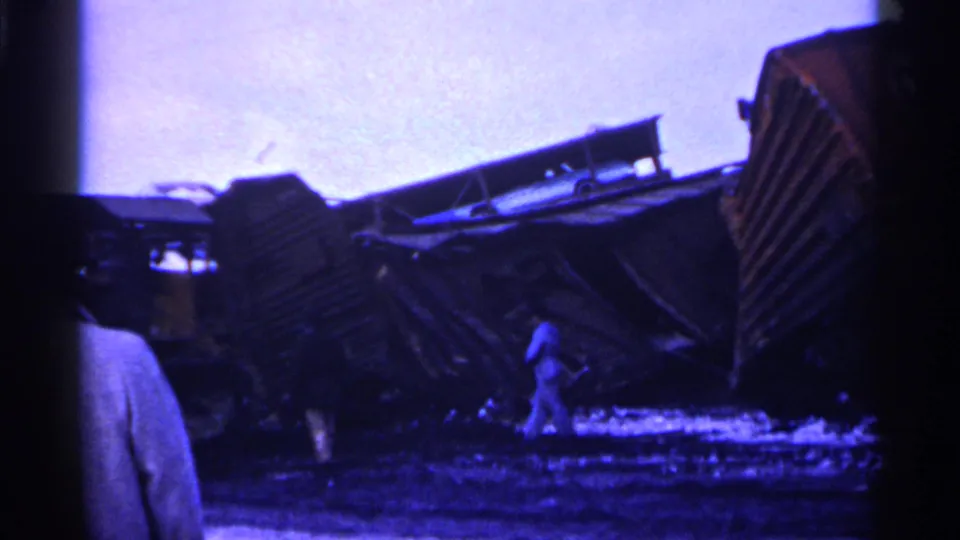 1961 Minnesota 8mm frame showing overturned freight cars, scattered debris, and people standing near a train derailment scene.