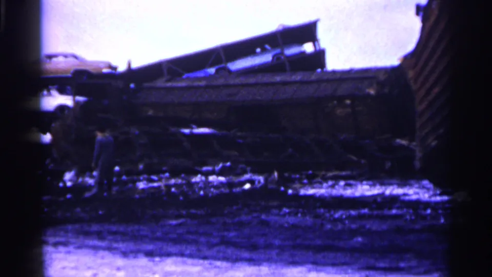 Minnesota 1961 home-movie frame with a person walking through the foreground of a train derailment aftermath.