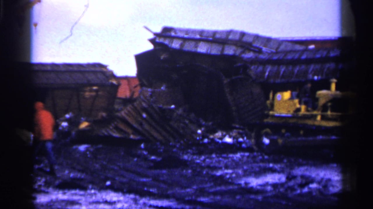 Wrecked rail car and debris in a muddy junkyard with a person in an orange jacket at left.