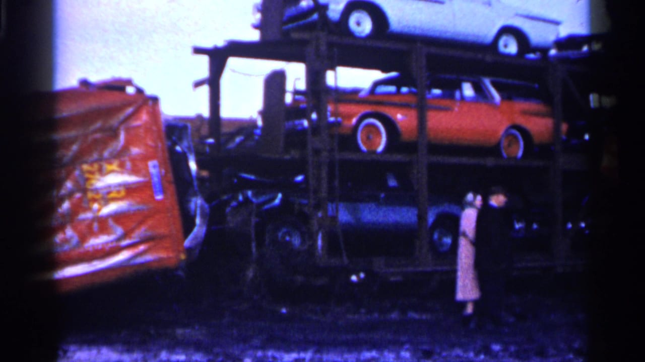 Stacked cars on a carrier beside a red-orange crushed panel, with two adults standing nearby.