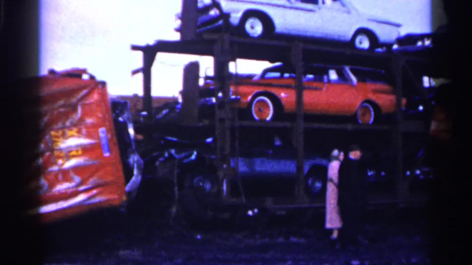 1961 Minnesota home-movie frame of stacked mid-century cars on a carrier with two adults standing in the junkyard foreground.