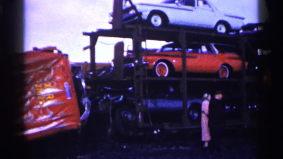 1961 Minnesota junkyard frame showing two adults beside a multi-level car carrier loaded with older automobiles.