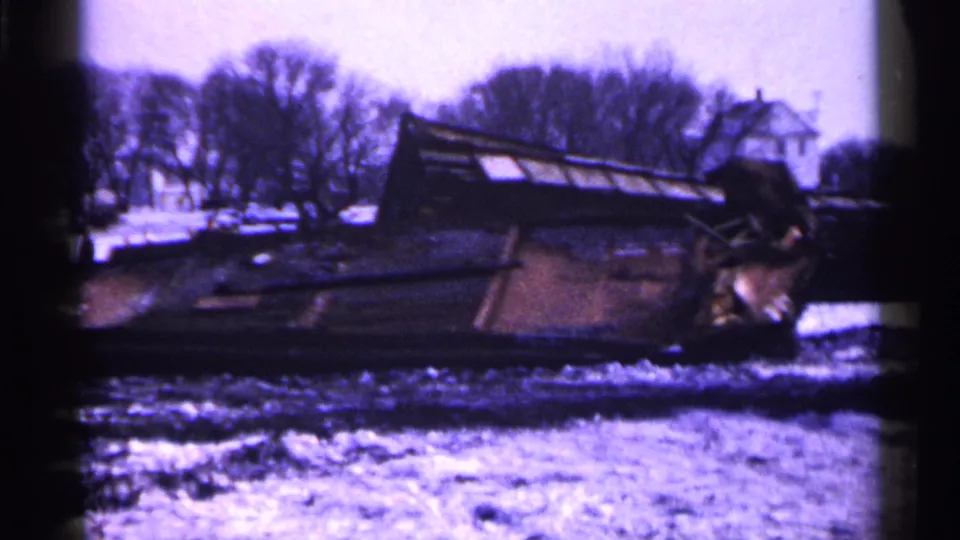 Minnesota 1961 frame of a derailed rail car on its side in snow, with bare trees and distant buildings in the background.