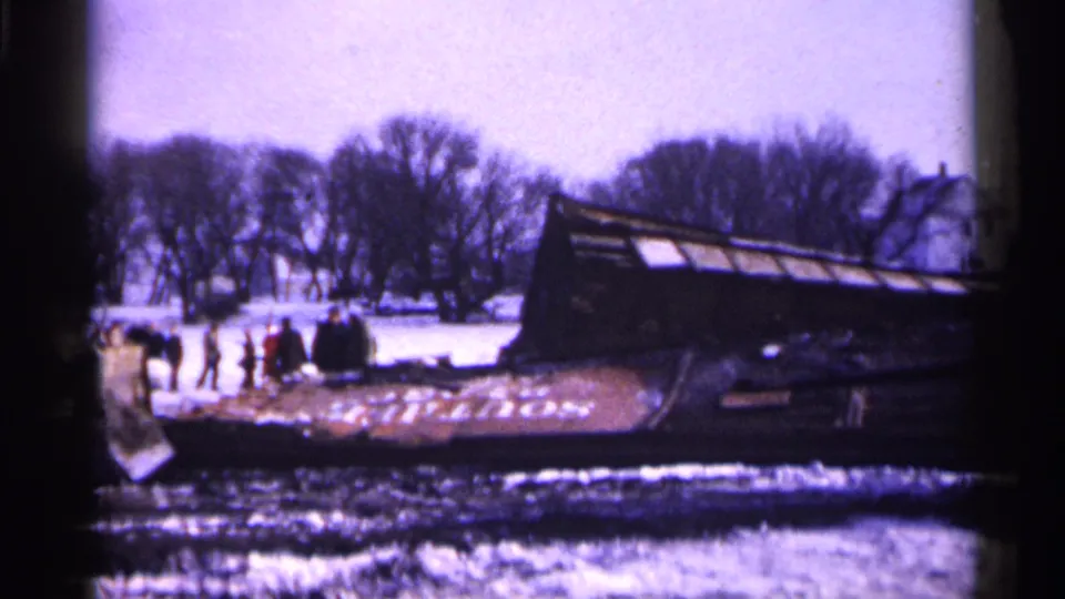 Minnesota 1961 frame showing people gathered near a rail car lying on its side, with snow, bare trees, and homes behind it.