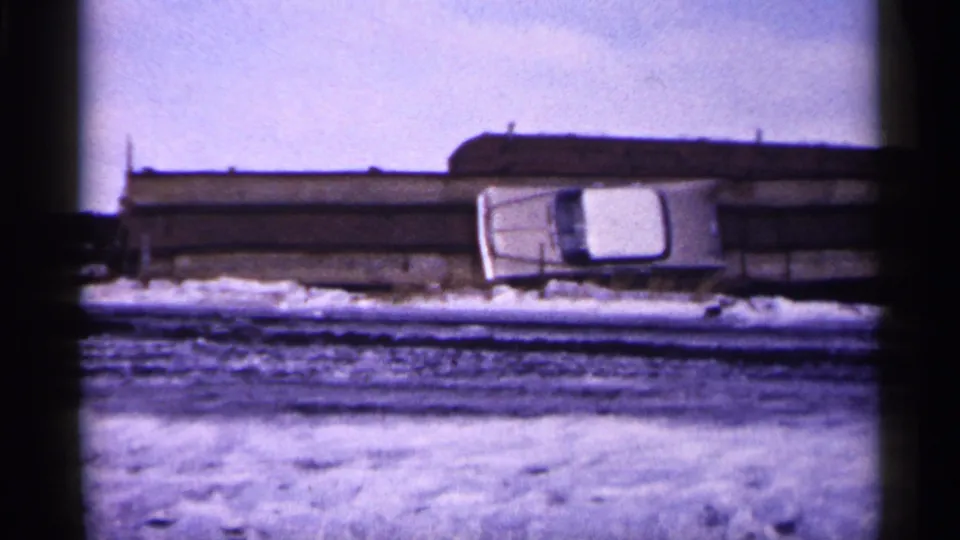 1961 Minnesota 8mm frame showing a white car tilted beside a bridge or barrier with snow and a long structure in the background.