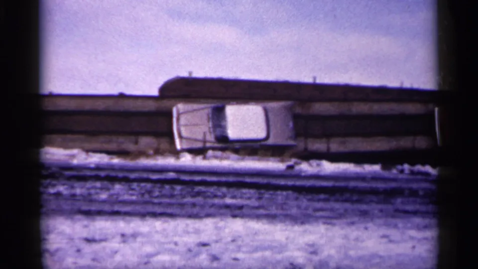 1961 Minnesota 8mm frame with a white car near a snowy bridge or roadside barrier, seen through soft grain and dark frame edges.