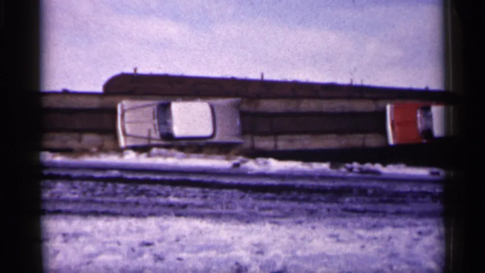 1961 Minnesota 8mm frame showing the white car by a snowy barrier as a red vehicle appears at the right edge of the scene.
