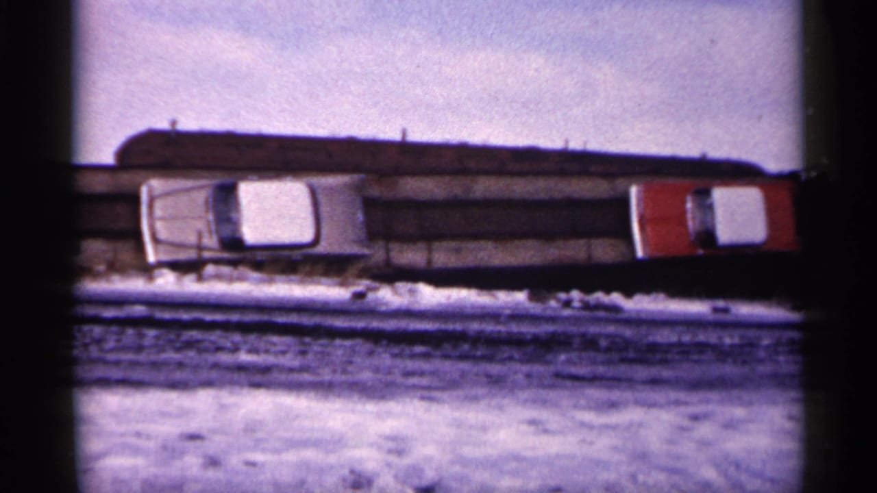 Snowy yard with a long dark scrap pile, a white car, and a red vehicle seen in soft 8mm color.