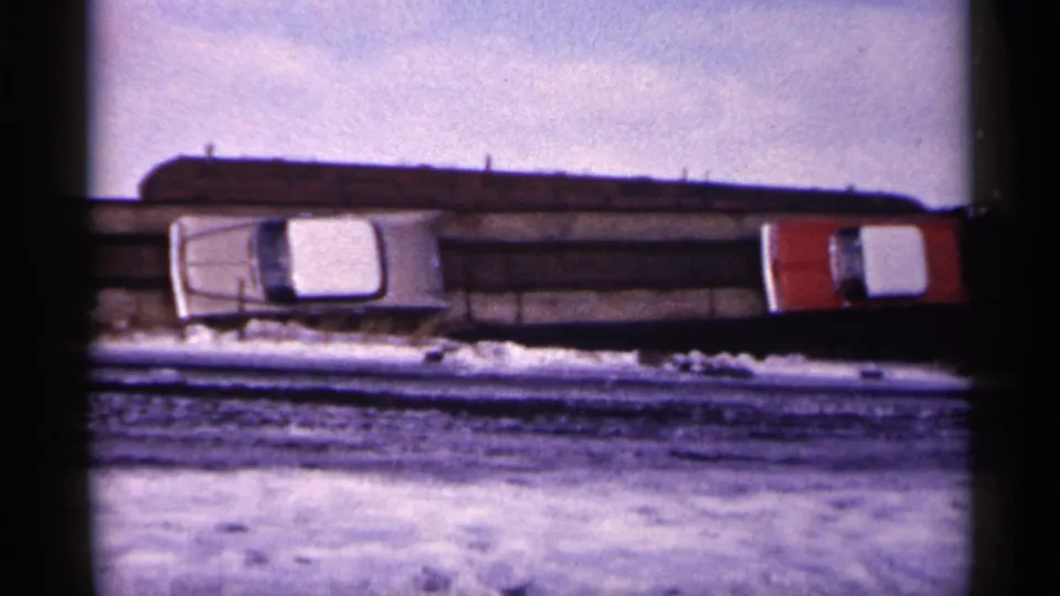 1961 Minnesota 8mm frame showing snow-covered ground, a white sedan, and a red vehicle against a dark junkyard pile.