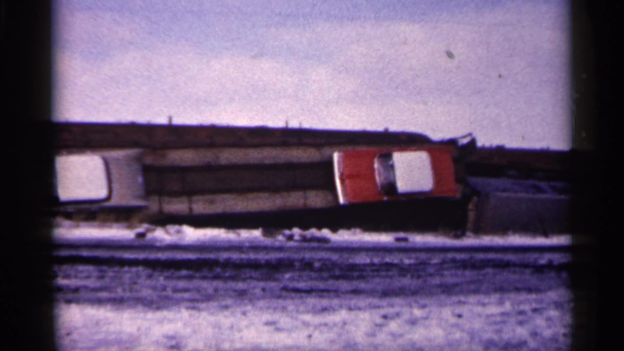 Distant snowy junkyard view with a white car, red vehicle, dark debris, and pale winter sky.