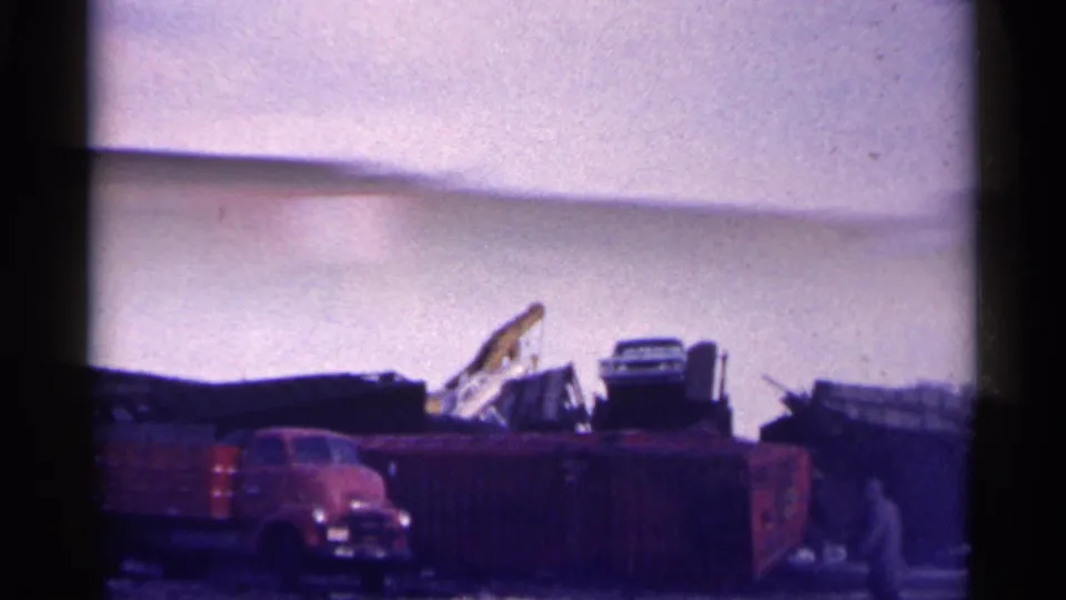 1961 Minnesota 8mm frame showing a red truck, yard debris, a yellow crane arm, and a distant man walking near the salvage area.