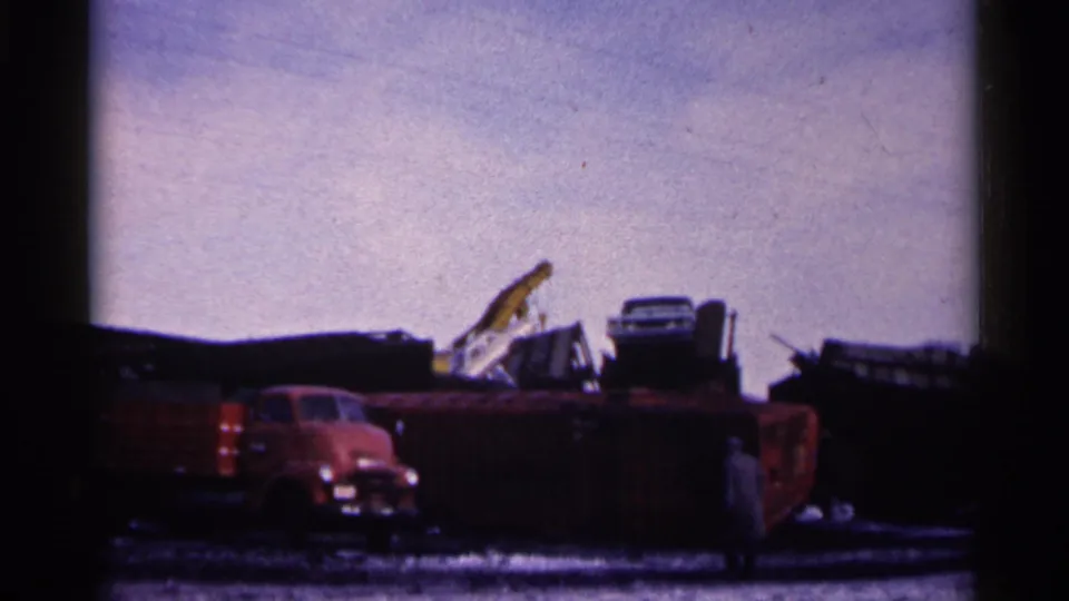 1961 Minnesota home-movie frame of a red truck in a snowy junkyard, with scrap piles, crane equipment, and a distant person visible.