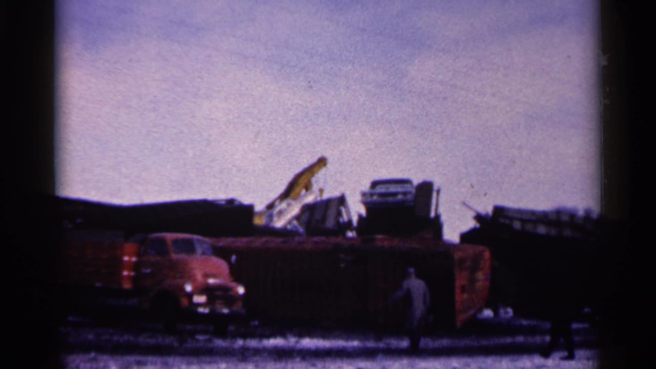 1961 Minnesota 8mm frame showing a red truck near piled scrap and wrecked vehicles, with distant people walking through the yard.