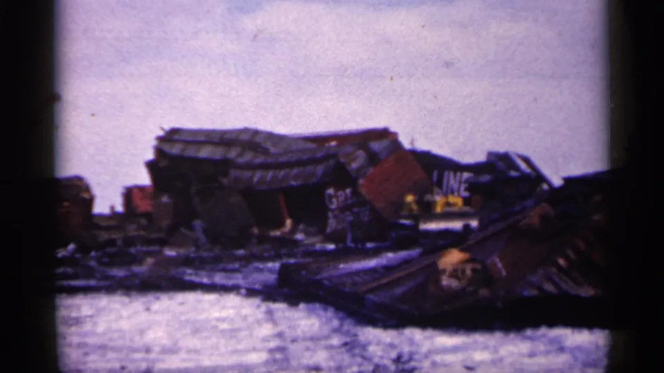 1961 Minnesota 8mm frame of junkyard debris, partial painted lettering, wrecked vehicles, and a distant person near the scrap.