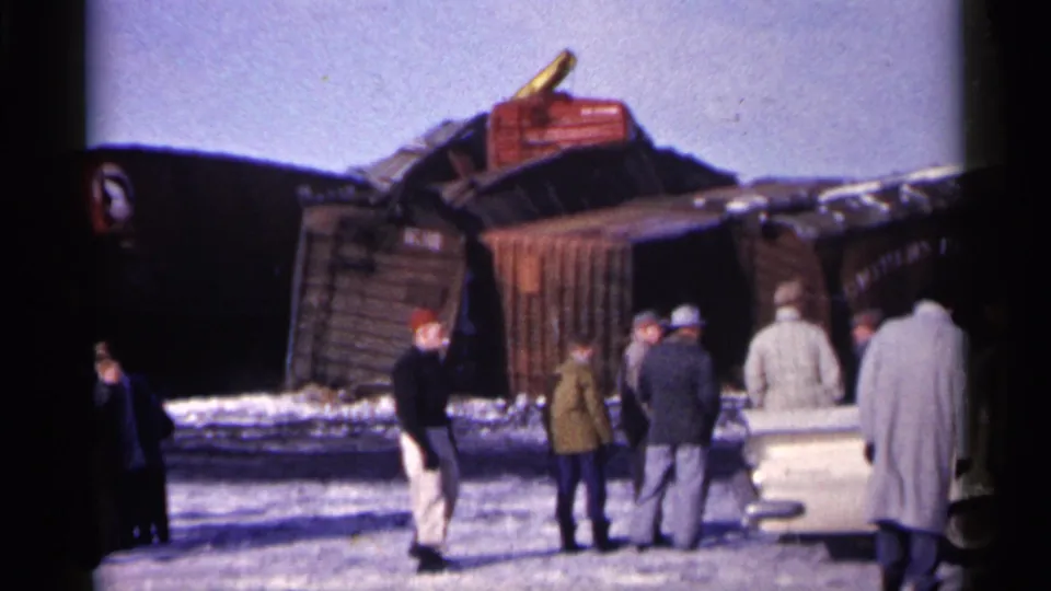 1961 Minnesota home-movie frame showing bundled onlookers and a period car beside a train derailment scene.