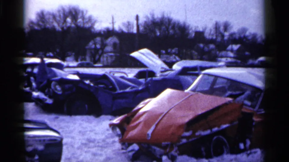 1961 Minnesota 8mm frame of a snowy salvage yard with damaged vintage automobiles and an orange car with a crushed front hood.