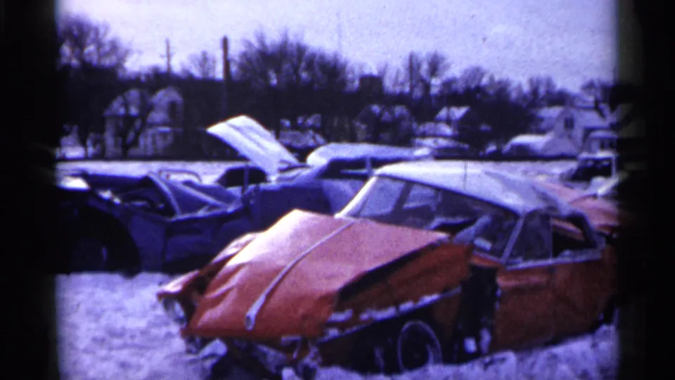 Snow-covered 1961 Minnesota salvage-lot view with damaged midcentury cars, a raised hood, bare trees, and houses in the background.