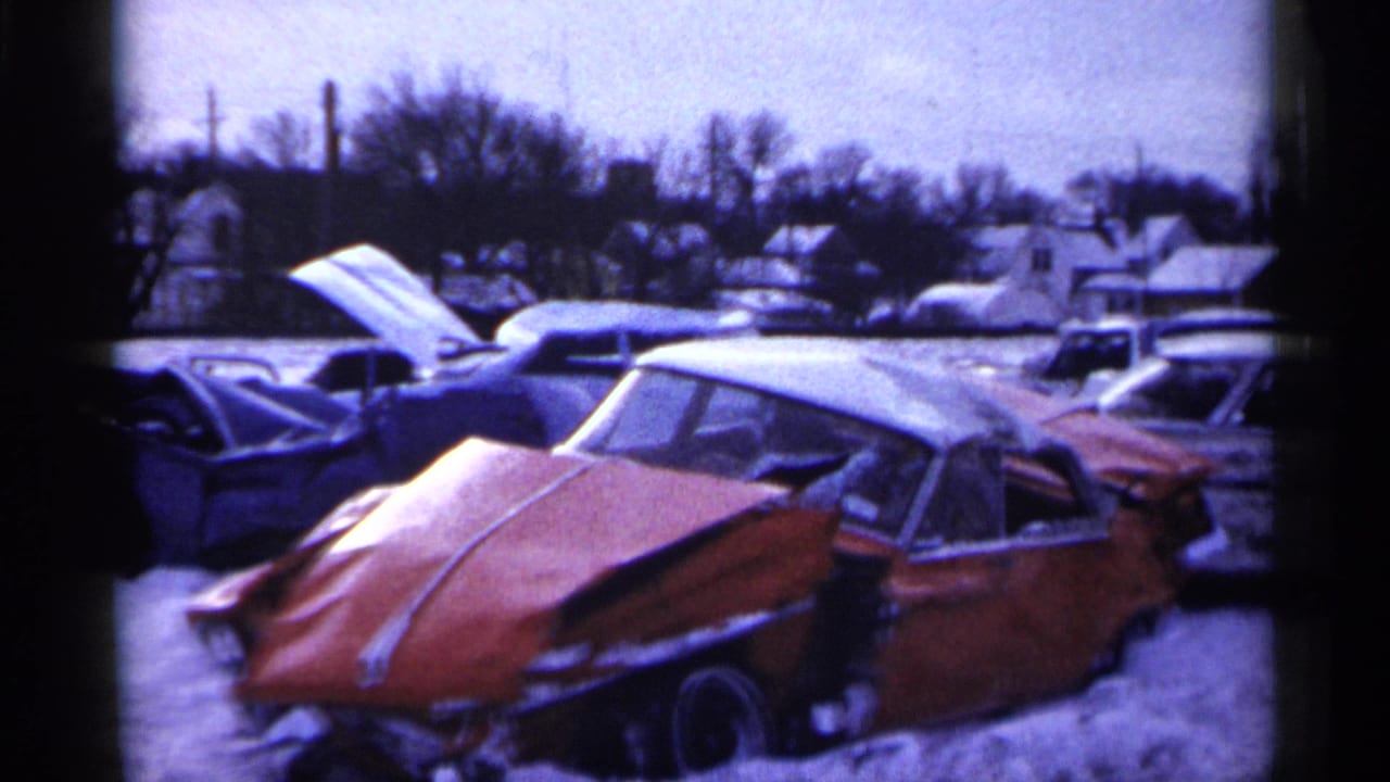 1961 Minnesota 8mm frame showing a snow-covered junkyard with a damaged red car in front and other mid-century cars behind it.
