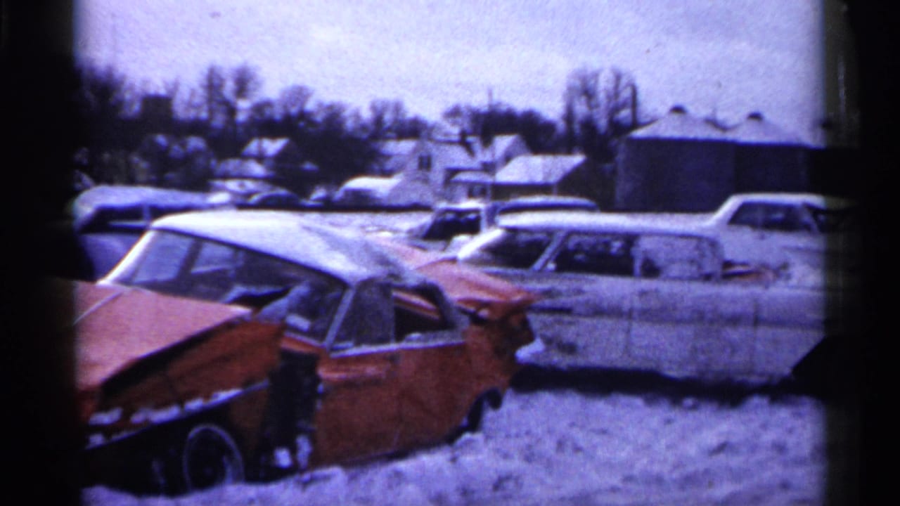 1961 Minnesota junkyard frame with a wrecked red car, snow on the ground, and several mid-century vehicles parked nearby.