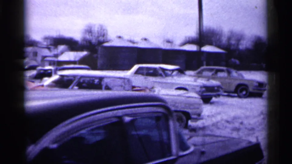 1961 Minnesota 8mm frame showing a snowy yard with several older cars, small buildings, bare trees, and a dark foreground vehicle roof.
