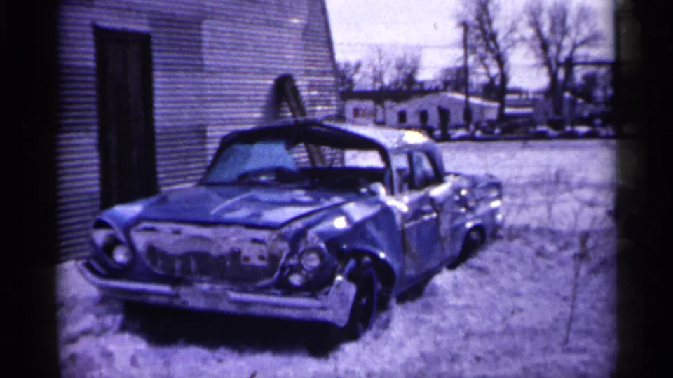 1961 Minnesota frame showing a damaged blue car parked beside a small building, with snow, other cars, and bare trees in the background.