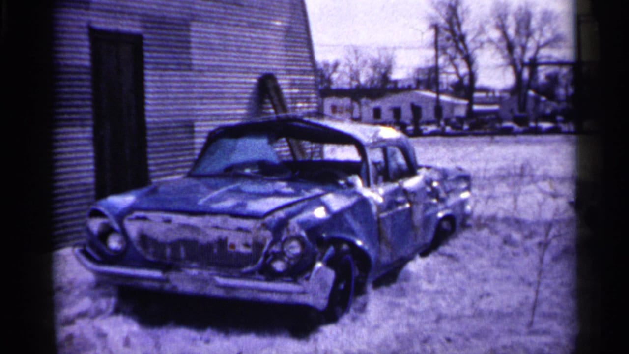 Damaged car parked in snow beside a building, with other vehicles and bare trees in the background.