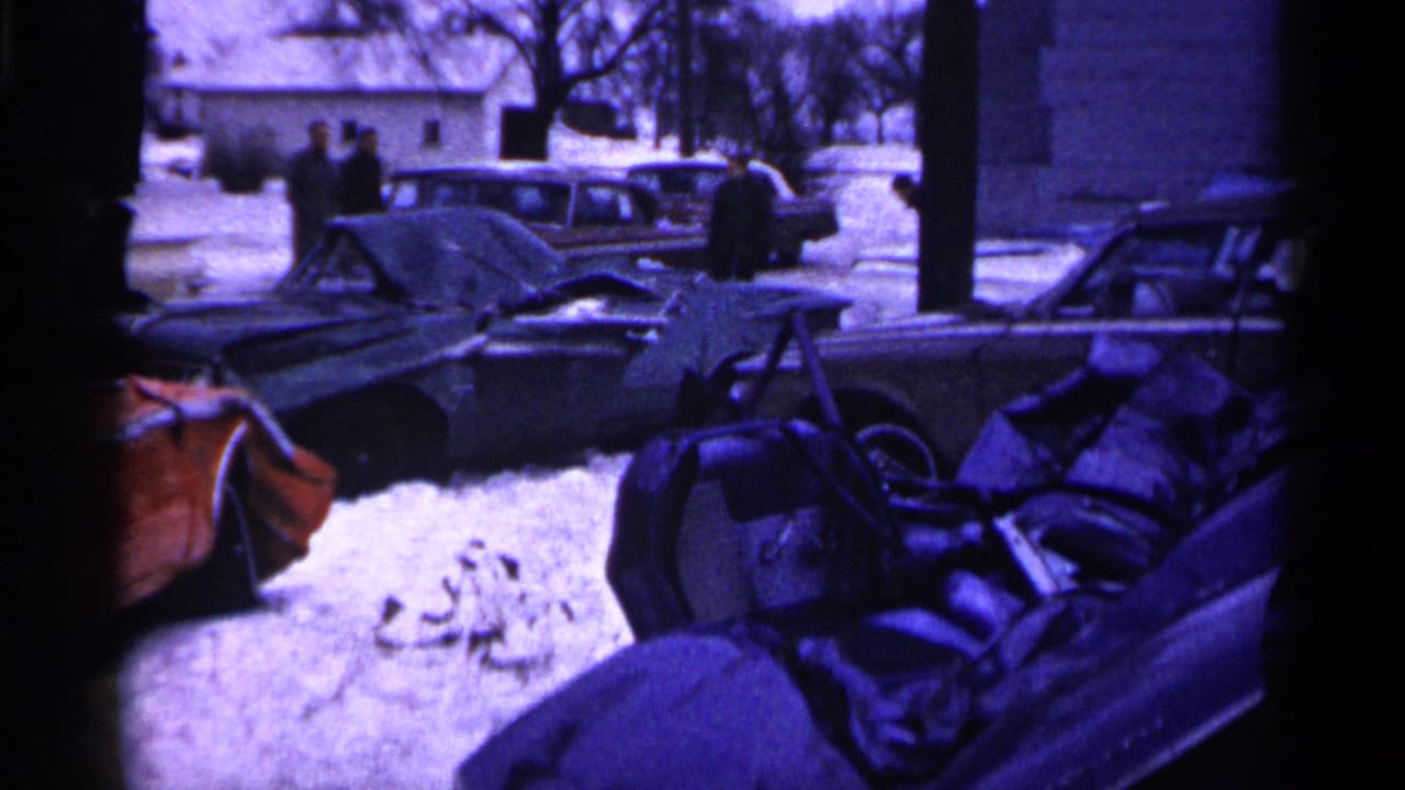 Damaged classic cars are parked in a snowy Minnesota lot as people in winter coats stand nearby in this 1961 home-movie frame.