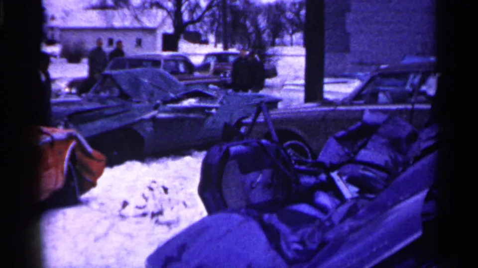 A snow-covered wrecked car with an open door sits in a Minnesota lot, with people and buildings visible in the background.