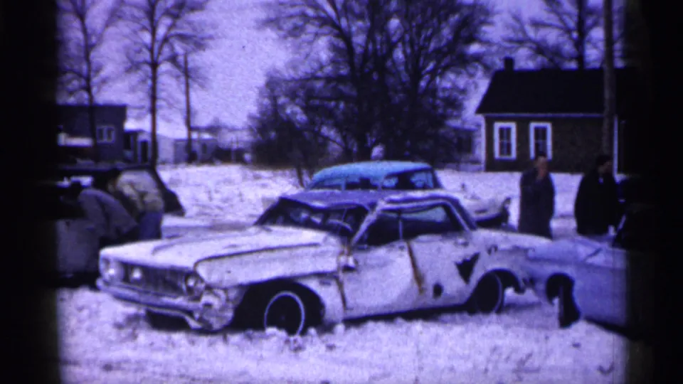 A white wrecked car sits low in the snow while people stand nearby in a 1961 Minnesota home-movie view.