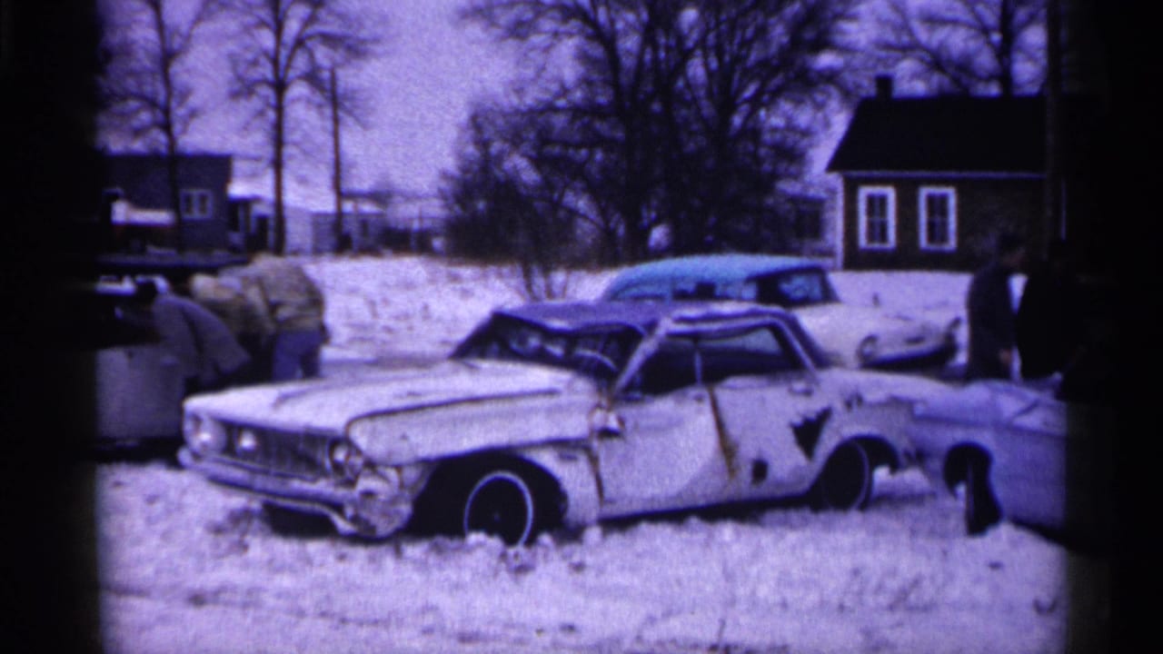 Minnesota 1961 home-movie frame showing a damaged American sedan in snow with bundled onlookers and houses in the background.