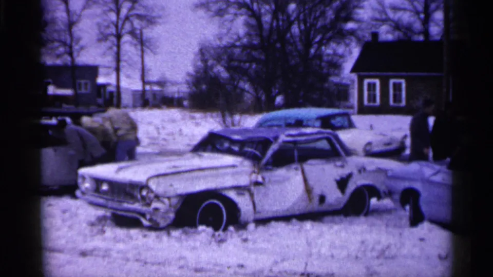 Minnesota 1961 home-movie frame showing a damaged American sedan in snow with bundled onlookers and houses in the background.