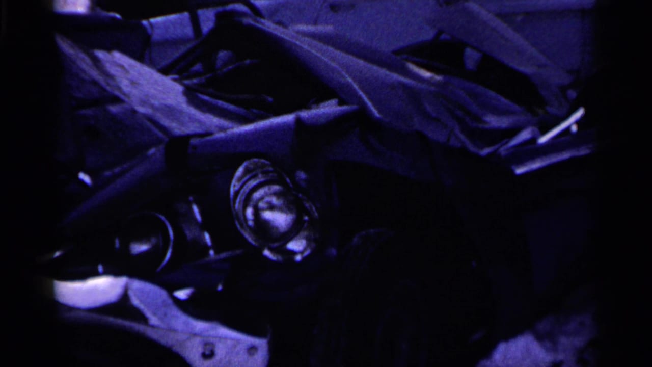 Close 8mm view of a wrecked car section, with a headlight, folded sheet metal, and strong purple color cast.
