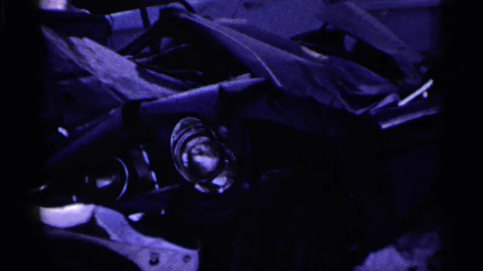 Close 8mm view of a wrecked car section, with a headlight, folded sheet metal, and strong purple color cast.
