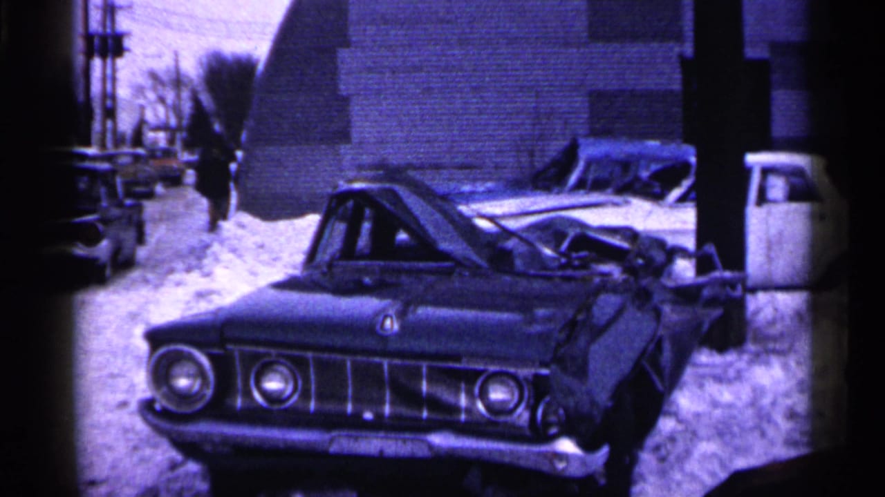 1961 Minnesota 8mm frame showing a crushed pale car in snow, with parked cars, brick buildings, and a distant pedestrian nearby.