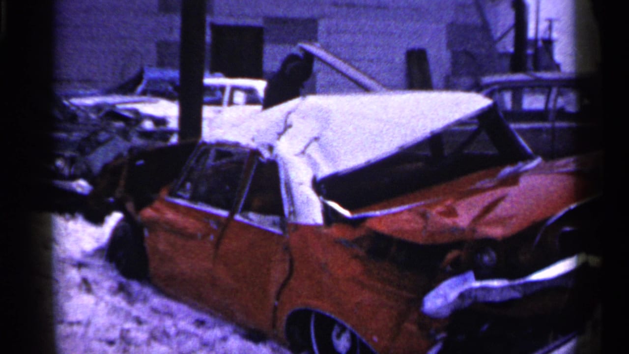 1961 Minnesota frame of a red car with severe rear damage in snow, near other wrecked cars and industrial-looking buildings.