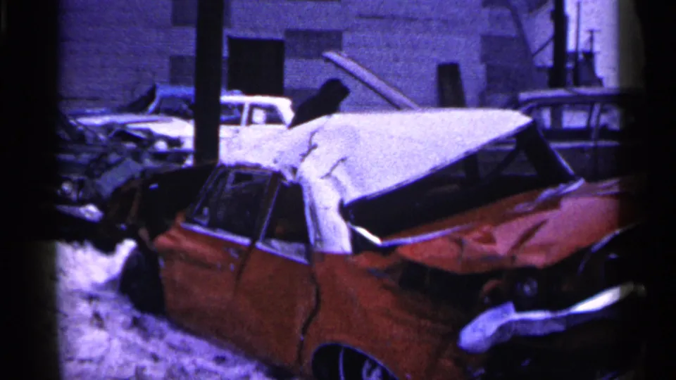 1961 Minnesota 8mm view of a red car with major body damage in snow, with other damaged vehicles and buildings behind it.