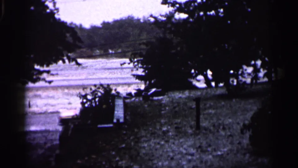1961 Minnesota 8mm view of a winter road beyond a yard, framed by trees, shrubs, and film gate shadows.