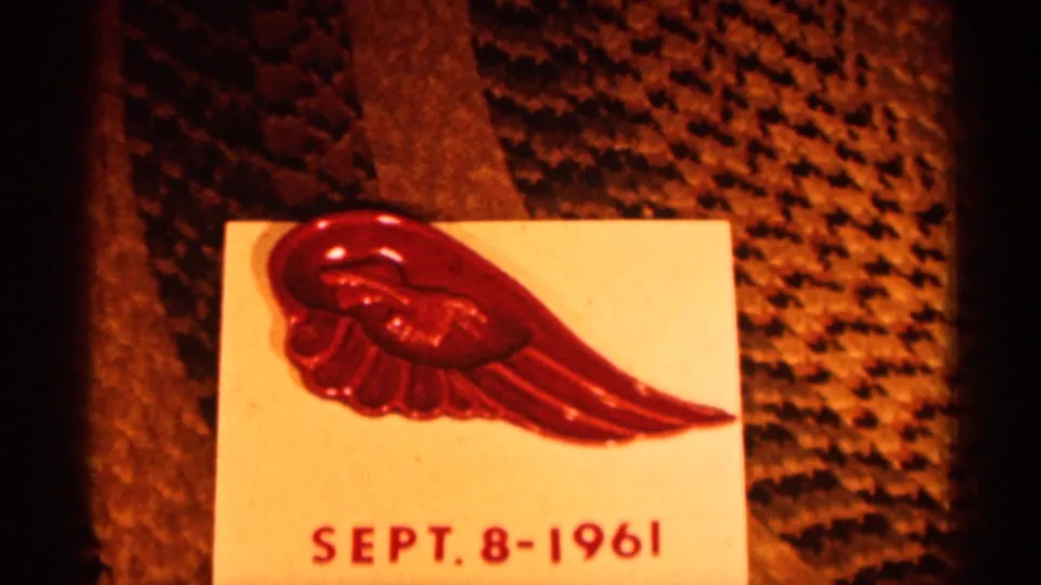 Close 1961 8mm frame of a red wing-shaped pin resting on a white card dated Sept. 8-1961 on textured fabric.