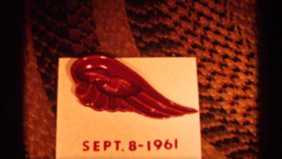 Close-up of a red wing-shaped pin on a dated card, with Sept. 8-1961 visible against patterned fabric.