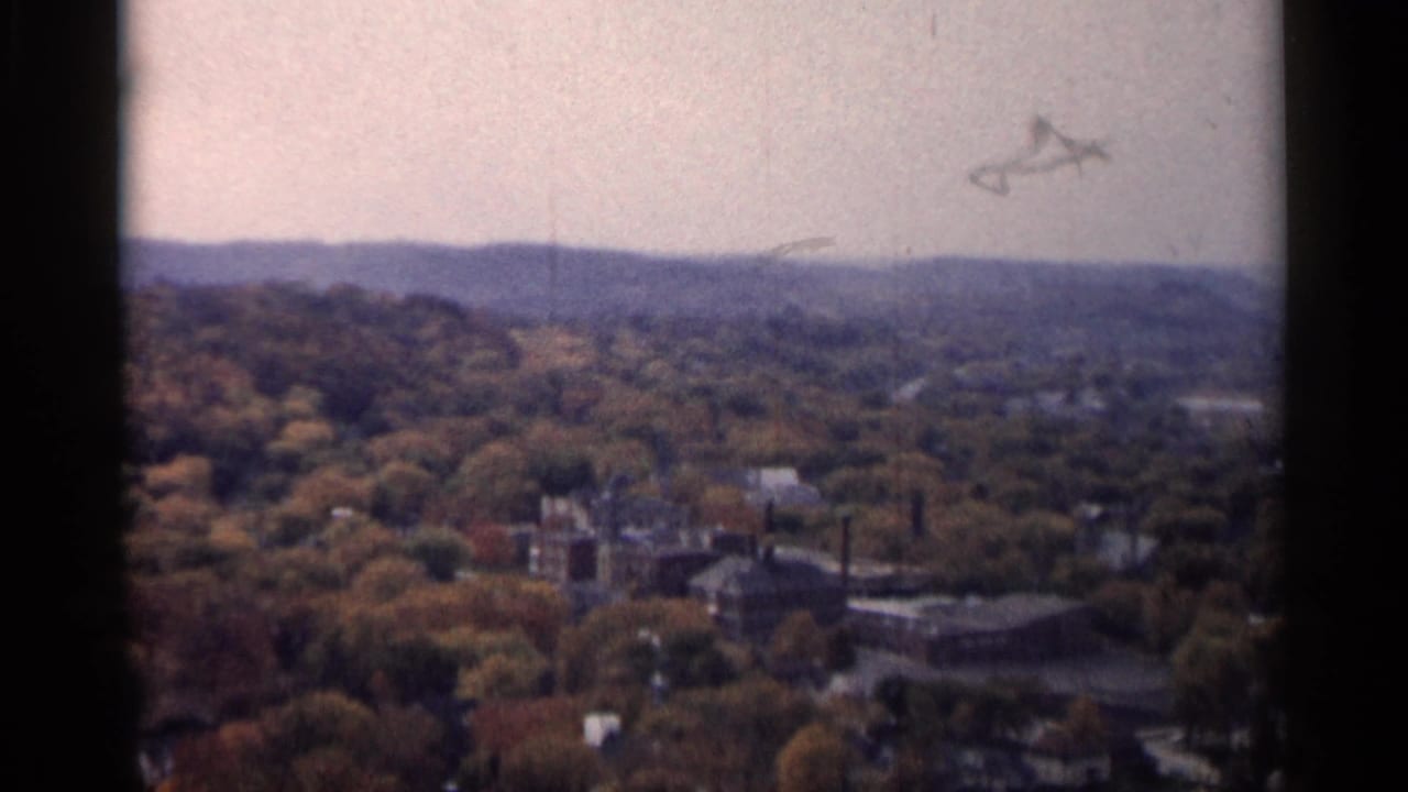 Soft aerial view over autumn trees, town buildings, hills, and an industrial complex.
