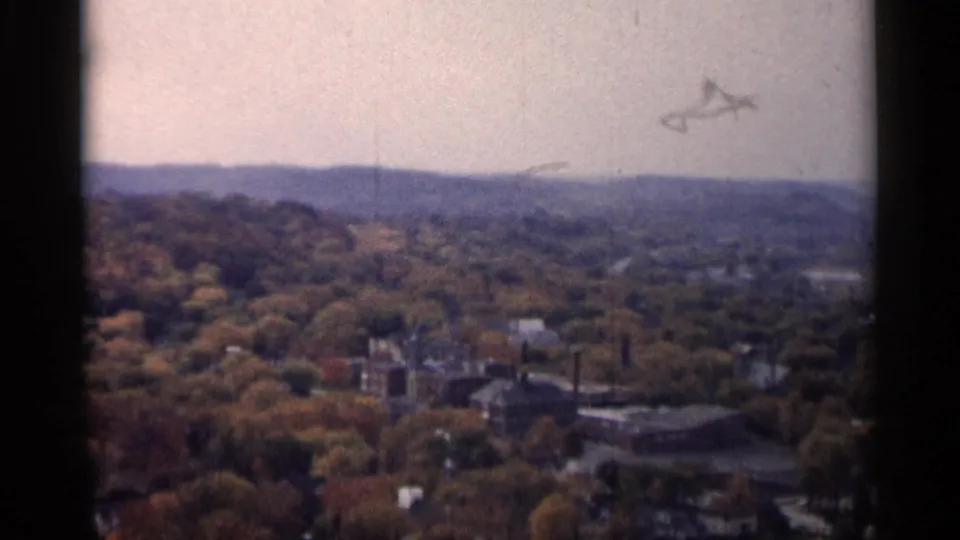 1961 Minnesota 8mm frame showing an aerial town view with autumn woodland, low buildings, distant hills, and industrial structures.