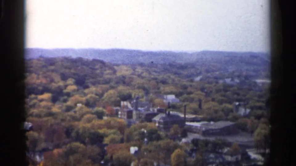 1961 Minnesota home-movie frame looking over autumn foliage, low buildings, industrial stacks, and a hazy hill line.