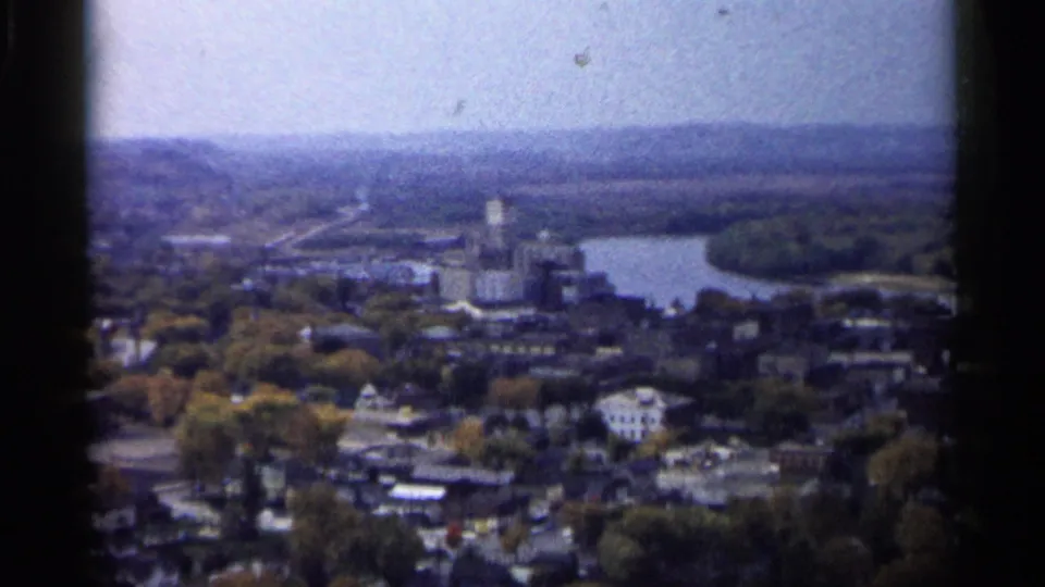 1961 Minnesota frame showing a lakeside town from above, with autumn trees, clustered buildings, roads, distant hills, and water.