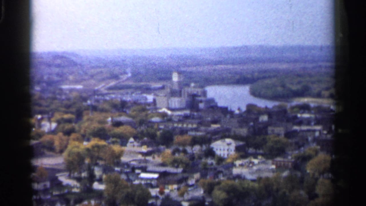 1961 Minnesota 8mm aerial view over a tree-filled town, with low buildings, a river bend, and hazy hills in the distance.