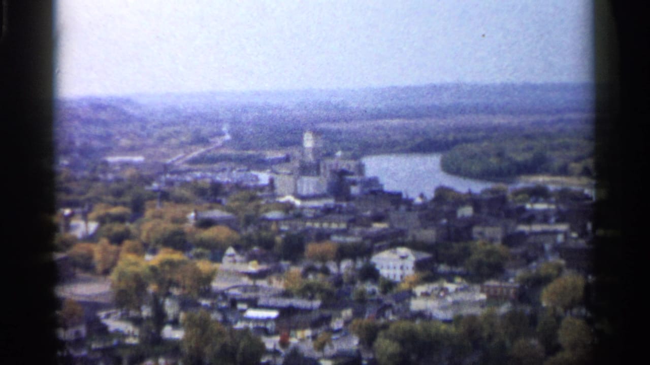 Aerial town scene with river, clustered buildings, trees, and horizon framed by film edges.