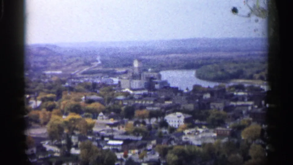 1961 Minnesota home-movie aerial view with a tall riverside structure, dense trees, town blocks, and distant wooded hills.