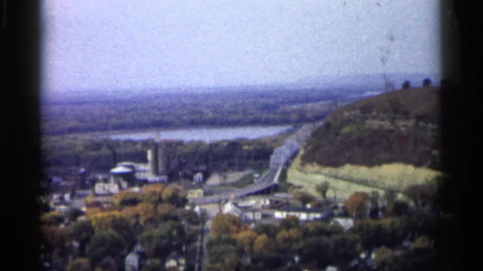 1961 Minnesota aerial frame showing a hillside above the town, a curving road, river water, trees, and scattered low buildings.