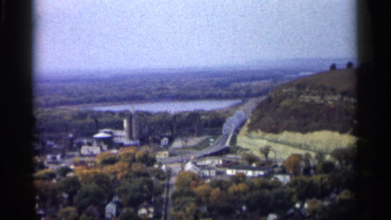 1961 Minnesota home-movie view of a river valley town with a bridge, tower-like structure, and sloping wooded land.