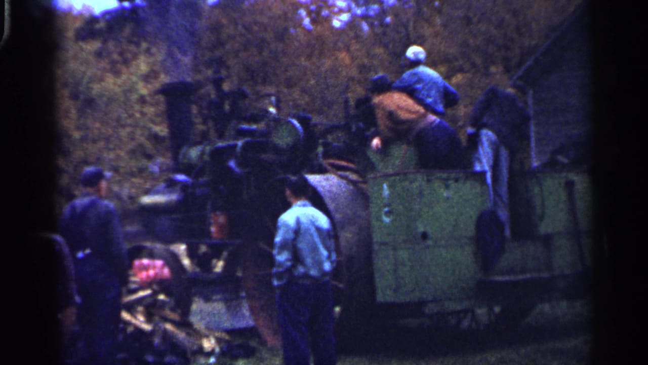 1961 Minnesota 8mm frame showing men in work clothes around a large green steam-powered agricultural machine outdoors.